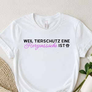 May include: White t-shirt with the German text "WEIL TIERSCHUTZ EINE Herzenssache IST" in black and pink lettering, along with a paw print. The shirt is styled with a knot at the bottom.