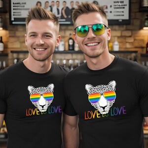Puede incluir: Dos personas con camisetas negras con un diseño de cabeza de leopardo. El leopardo lleva gafas de sol arcoíris, y el texto "LOVE is LOVE" está debajo en colores arcoíris. El fondo es un bar.