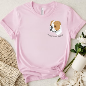 Könnte beinhalten: Hellrosa T-Shirt mit einem Bulldoggen-Motiv und dem Text "Adopt. Love. Repeat." Der Bulldogge ist in Braun und Weiß mit schwarzer Umrandung dargestellt. Das T-Shirt besteht aus einem weichen Material und hat einen Rundhalsausschnitt.