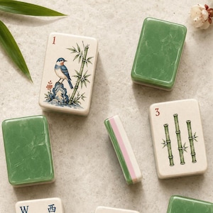 May include: A collection of rectangular mahjong tiles featuring various designs including a bird on a rock, bamboo stalks, a bridge, and floral patterns. Some tiles have solid green backs, while others display numbers and symbols in a white finish.