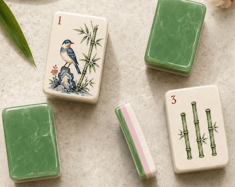 May include: A collection of rectangular mahjong tiles featuring various designs including a bird on a rock, bamboo stalks, a bridge, and floral patterns. Some tiles have solid green backs, while others display numbers and symbols in a white finish.