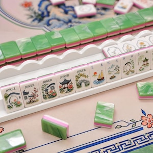 Pode incluir: Um conjunto de pe&ccedil;as de Mahjong com versos marmorizados em verde e branco sobre uma camada rosa. As faces frontais exibem ilustra&ccedil;&otilde;es coloridas de paisagens, pagodes e pontes, com algumas pe&ccedil;as marcadas como Joker, E, N e W em preto.