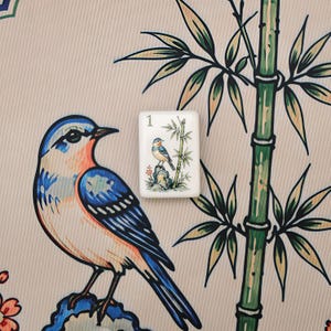 Pode incluir: Uma pe&ccedil;a retangular branca de mahjong com o n&uacute;mero 1 e uma pequena ilustra&ccedil;&atilde;o de um p&aacute;ssaro azul pousado numa rocha ao lado de caules de bambu, colocada sobre um tecido com uma estampa maior de p&aacute;ssaro e bambu a condizer.