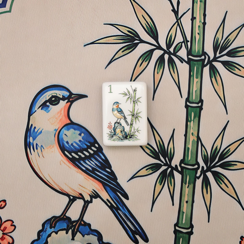 Pode incluir: Uma pe&ccedil;a retangular branca de mahjong com o n&uacute;mero 1 e uma pequena ilustra&ccedil;&atilde;o de um p&aacute;ssaro azul pousado numa rocha ao lado de caules de bambu, colocada sobre um tecido com uma estampa maior de p&aacute;ssaro e bambu a condizer.