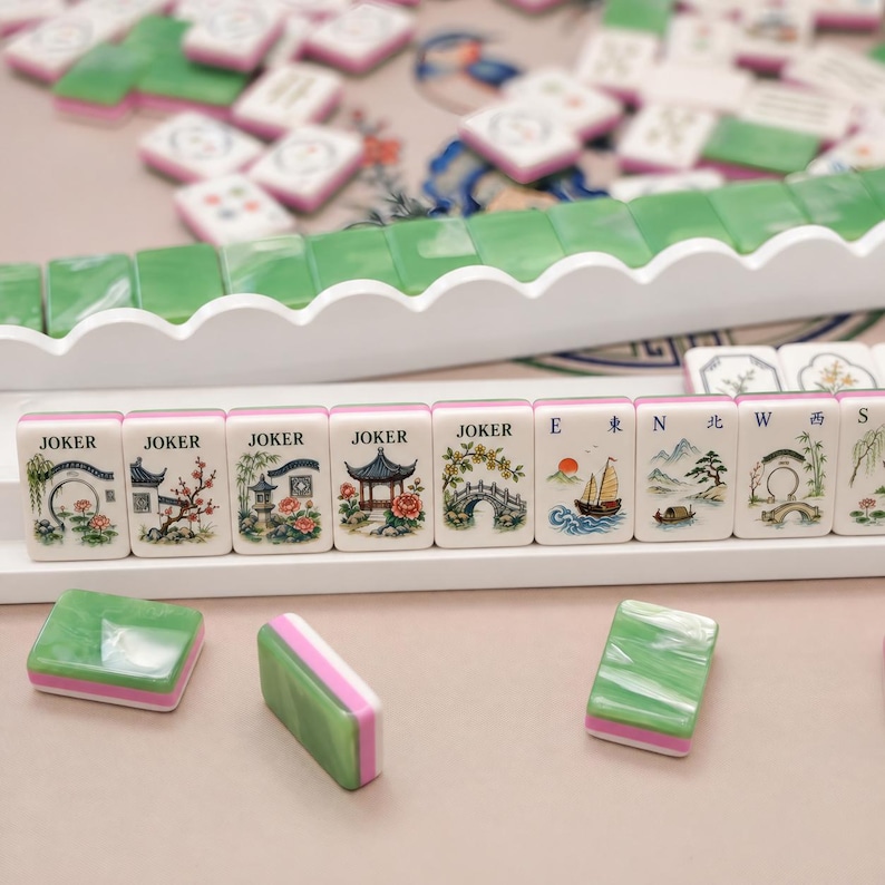 Pode incluir: Um conjunto de pe&ccedil;as de Mahjong com faces brancas contendo ilustra&ccedil;&otilde;es coloridas de paisagens, flores e a palavra JOKER, com versos verdes e rosas. As pe&ccedil;as est&atilde;o dispostas em suportes brancos com outras pe&ccedil;as soltas por perto.