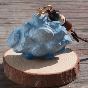 Puede incluir: Un llavero de madera azul tallado a mano con forma de nube. El llavero tiene un anillo dorado y una borla decorativa de cuentas. La nube se encuentra sobre una pequeña rodaja de madera redonda con borde de corteza natural.