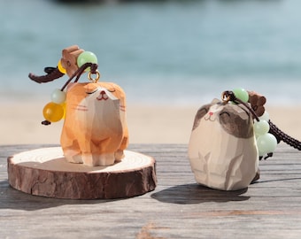 May include: Two wooden cat figurines with keychains. One is orange and white, the other is white and brown. Both have closed eyes and are smiling. They are on a wooden surface with a blurred background of the ocean.