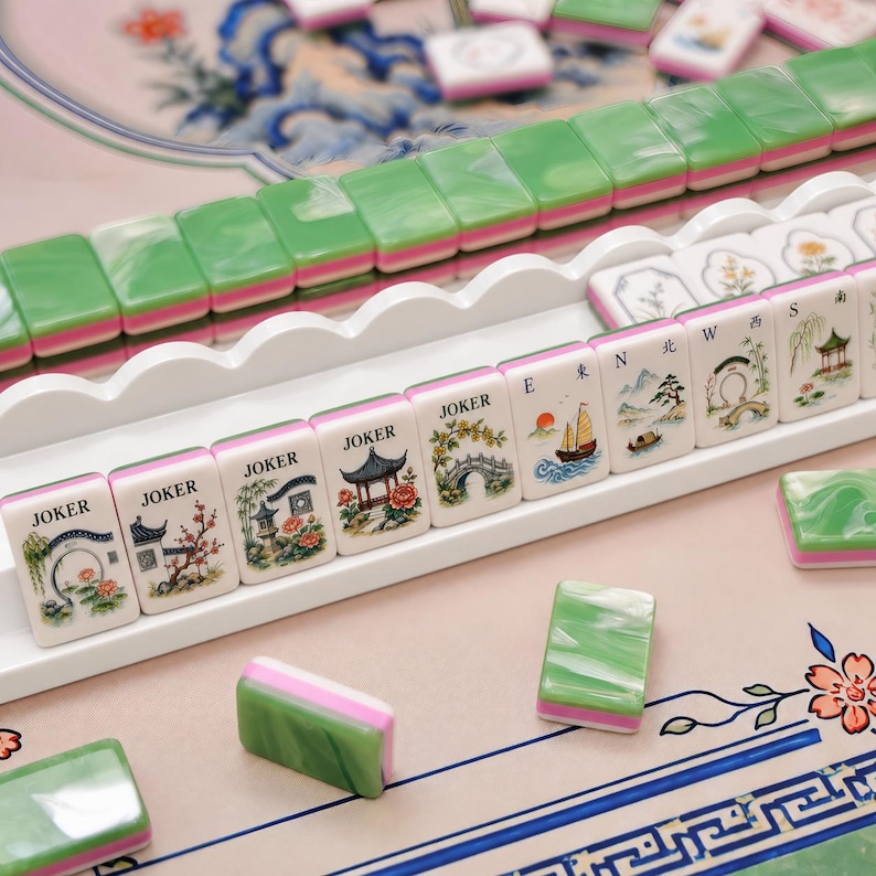 Puede incluir: Un juego de fichas de Mahjong con dorsos veteados en verde y blanco sobre una capa rosa. Las caras frontales muestran ilustraciones coloridas de paisajes, pagodas y puentes, con algunas fichas marcadas como Joker, E, N y W en negro.