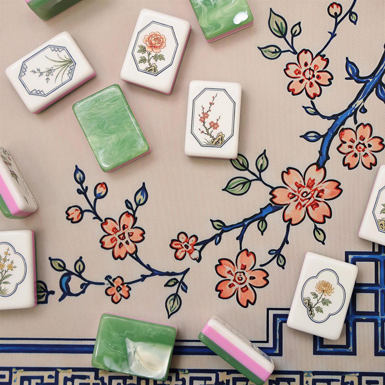 Pode incluir: Uma cole&ccedil;&atilde;o de pe&ccedil;as retangulares de mahjong espalhadas sobre uma superf&iacute;cie bege com um padr&atilde;o estampado de ramos azuis e flores de cerejeira cor-de-rosa. As pe&ccedil;as s&atilde;o brancas com o verso verde e apresentam ilustra&ccedil;&otilde;es florais.