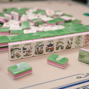 Pode incluir: Um conjunto de pe&ccedil;as retangulares de Mahjong com superf&iacute;cies marmorizadas em verde e branco e bases cor-de-rosa. V&aacute;rias pe&ccedil;as est&atilde;o alinhadas, exibindo ilustra&ccedil;&otilde;es de pagodes, pontes, p&aacute;ssaros e bambu, com o texto JOKER, BAM, CRAK.