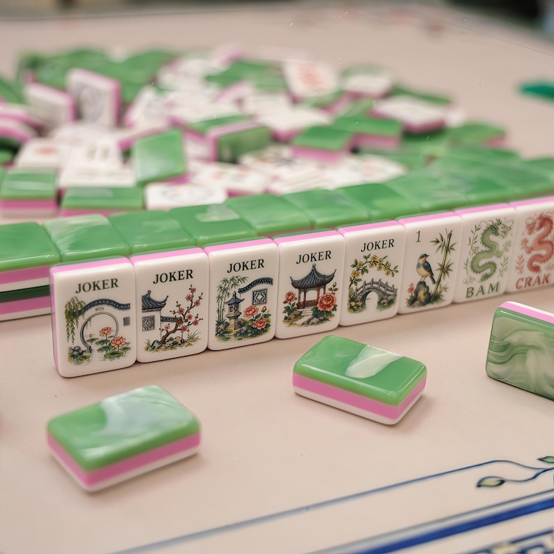 Pode incluir: Um conjunto de pe&ccedil;as retangulares de Mahjong com superf&iacute;cies marmorizadas em verde e branco e bases cor-de-rosa. V&aacute;rias pe&ccedil;as est&atilde;o alinhadas, exibindo ilustra&ccedil;&otilde;es de pagodes, pontes, p&aacute;ssaros e bambu, com o texto JOKER, BAM, CRAK.