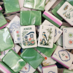 May include: A collection of mahjong tiles featuring a mix of solid green marbled tiles and white tiles with detailed illustrations. One tile displays the word JOKER above a garden scene, while another shows a bird perched on a bamboo stalk.