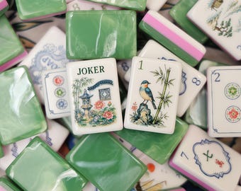 Könnte beinhalten: Eine Sammlung von Mahjong-Steinen, bestehend aus einer Mischung aus einfarbig grünen, marmorierten Steinen und weißen Steinen mit detaillierten Illustrationen. Ein Stein zeigt das Wort JOKER über einer Gartenszene, ein anderer einen Vogel.