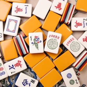 Puede incluir: Una colección de fichas de Mahjong rectangulares con una mezcla de reversos de color naranja sólido y caras blancas. Las fichas blancas presentan varios diseños, incluyendo caracteres chinos rojos, ilustraciones de aves verdes, círculos azules y verdes y la palabra JOKER.