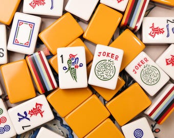 Pode incluir: Uma coleção de peças retangulares de Mahjong com uma mistura de versos laranja sólidos e faces brancas. As peças brancas apresentam vários desenhos, incluindo caracteres chineses vermelhos, ilustrações de pássaros verdes, círculos azuis e verdes e a palavra JOKER.