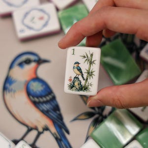 Pode incluir: Uma pe&ccedil;a retangular branca de mahjong com o n&uacute;mero 1 e uma ilustra&ccedil;&atilde;o detalhada de um pequeno p&aacute;ssaro azul pousado numa rocha ao lado de um caule de bambu verde. A pe&ccedil;a &eacute; segurada contra um fundo com um p&aacute;ssaro azul ilustrado.