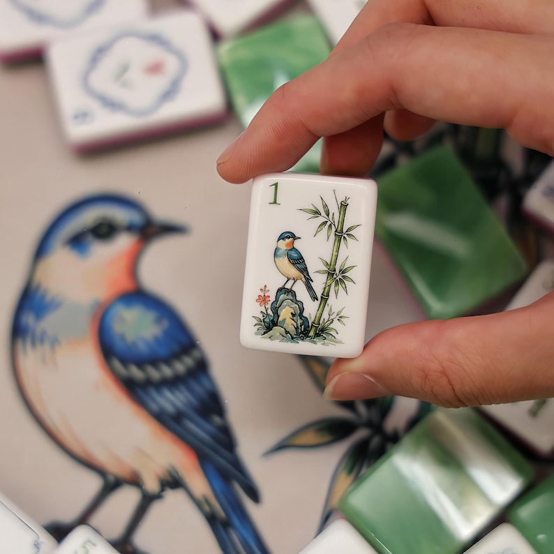 Puede incluir: Una ficha de Mahjong rectangular blanca con el n&uacute;mero 1 en verde en la parte superior, que muestra una ilustraci&oacute;n detallada de un peque&ntilde;o p&aacute;jaro azul y naranja posado sobre una roca junto a un tallo de bamb&uacute; verde, sobre otras fichas.