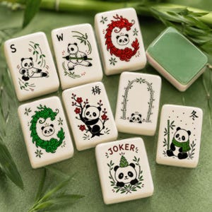 May include: A set of white rectangular mahjong tiles featuring various illustrations of pandas. Designs include pandas with bamboo, a red dragon, a plum blossom branch, a bamboo arch, and a tile labeled JOKER with a panda wearing a bamboo hat.