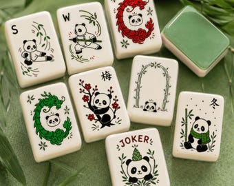 Puede incluir: Un conjunto de fichas de mahjong rectangulares blancas con varias ilustraciones de pandas. Los diseños incluyen pandas con bambú, un dragón rojo, una rama de ciruelo, un arco de bambú y una ficha JOKER con un panda con sombrero.