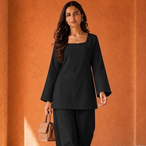 May include: A black long-sleeved tunic with a square neckline paired with matching black wide-leg trousers. The outfit is accessorized with a small tan structured handbag and open-toe sandals, creating a coordinated monochromatic ensemble.