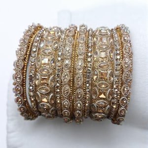 Peut inclure: Un ensemble de bracelets dorés, ornés de pierres précieuses incrustées et de motifs détaillés. Les bracelets sont empilés, présentant diverses textures et formes, reflétant la lumière et créant un aspect luxueux. Les bijoux sont posés sur un fond blanc.
