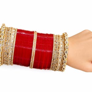 Può includere: Un set di braccialetti impilati con fasce cilindriche rosso scuro intervallate da polsini in metallo dorato. I polsini esterni sono decorati con pietre trasparenti sfaccettate in motivi a goccia e quadrati, creando un ornamento ampio.
