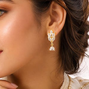 Puede incluir: Pendientes colgantes dorados con un diseño en forma de hoja, adornados con cristales claros y brillantes. Los pendientes presentan un grupo de cristales en la parte superior y un cristal triangular colgante en la parte inferior, creando un accesorio elegante.