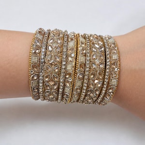 May include: A collection of gold-toned bangles, adorned with intricate floral and geometric patterns, encrusted with sparkling rhinestones and small pearls. The bangles are stacked on a wrist, showcasing their ornate design and elegant appeal.