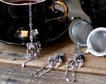 Puede incluir: Juego de tetera y taza de té negros con un infusor de té de calavera y huesos. La taza de té y la tetera son negras con detalles dorados. El infusor de té es un amuleto de calavera y huesos de plata en una cadena.