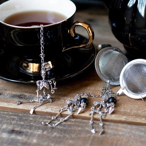 May include: Black teacup and teapot set with a skull and bones tea infuser. The teacup and teapot are black with gold accents. The tea infuser is a silver skull and bones charm on a chain.