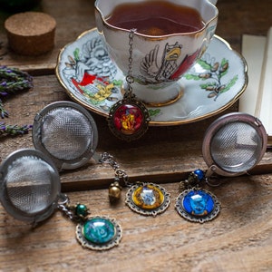 May include: A white teacup and saucer with a gold trim and a floral design. The teacup is filled with dark tea and has a tea infuser attached to the side. There are four tea infusers with colorful enamel charms on a wooden table.