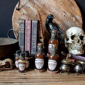 May include: A still-life arrangement featuring vintage books, a raven figurine, a skull, and several bottles labeled "PICKLED EARTHWORMS." The bottles have ornate metal tops and are displayed on a wooden surface, evoking a gothic or Halloween theme.