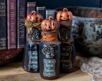 May include: Three decorative Halloween bottles with pumpkin-shaped stoppers. The bottles are black with labels that read "Spirits of Halloween." Each bottle is adorned with a decorative metal piece and wrapped with burlap and orange ribbon. The background includes antique books.