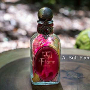 Puede incluir: Una botella de vidrio con una etiqueta dorada y roja que dice "Red Flame of the Bull". La botella est&aacute; llena de p&eacute;talos rosas y dorados y tiene un tap&oacute;n dorado y rojo.