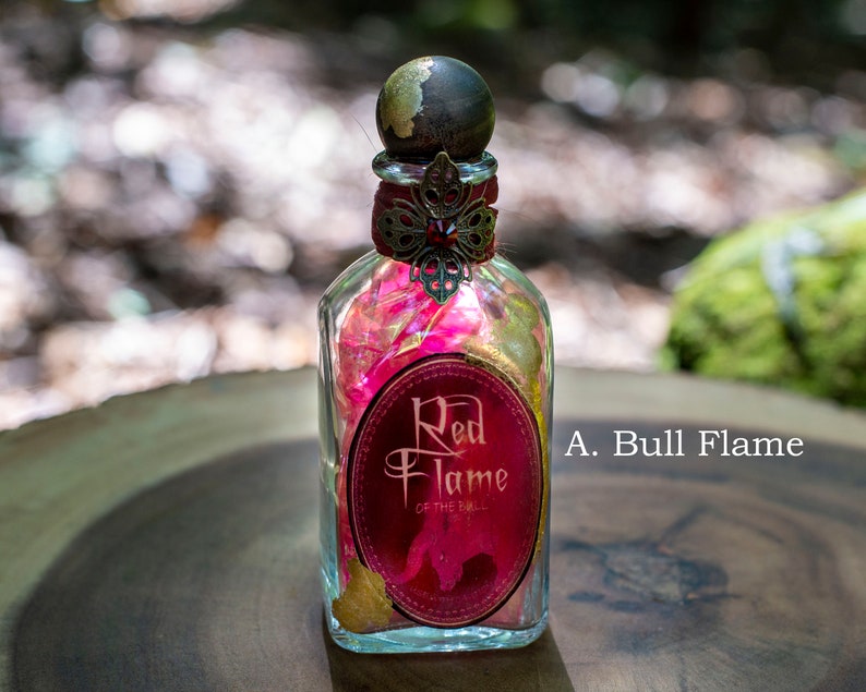 Puede incluir: Una botella de vidrio con una etiqueta dorada y roja que dice "Red Flame of the Bull". La botella est&aacute; llena de p&eacute;talos rosas y dorados y tiene un tap&oacute;n dorado y rojo.