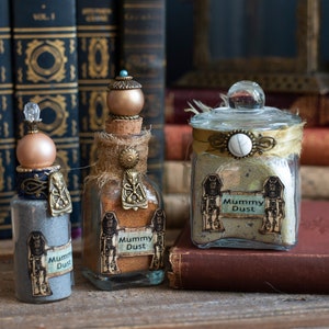 May include: Three glass jars with cork stoppers and decorative labels. The jars are filled with a brown powder and labeled "Mummy Dust". The jars are sitting on a wooden surface in front of a stack of books.