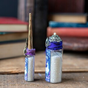 Puede incluir: Dos peque&ntilde;as botellas de vidrio llenas de purpurina blanca. Las botellas est&aacute;n decoradas con cinta morada y abalorios plateados. La botella de la izquierda tiene un cuerno de unicornio dorado en la parte superior. La botella de la derecha tiene una corona plateada en la parte superior.