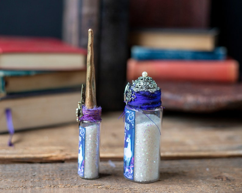 Puede incluir: Dos peque&ntilde;as botellas de vidrio llenas de purpurina blanca. Las botellas est&aacute;n decoradas con cinta morada y abalorios plateados. La botella de la izquierda tiene un cuerno de unicornio dorado en la parte superior. La botella de la derecha tiene una corona plateada en la parte superior.