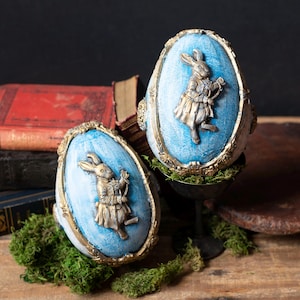 May include: Two decorative Easter egg ornaments. Each egg features a light blue oval center with a bronze-colored rabbit figure. The eggs are framed with ornate gold trim and set on a bed of green moss. The background includes antique books.