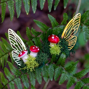 May include: Two gold fairy wings with red and white mushroom accents on a bed of green moss.