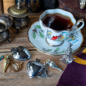 May include: A white teacup with a colorful design and a matching saucer, filled with dark tea. A silver tea infuser hangs from the cup, with a round, clear glass charm at the end. There are two silver heart-shaped tea infusers, a gold-colored bottle charm, and a purple and gold scarf in the background.