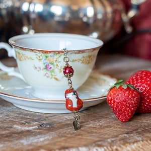 May include: A vintage teacup and saucer set with floral designs, a decorative tea infuser, and two fresh strawberries. The teacup is cream-colored with gold trim and a delicate floral pattern. The infuser features a red fox charm.