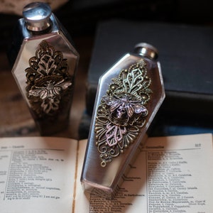 May include: Two silver metal hip flasks with ornate designs. The flasks are shaped like coffins and feature a butterfly design with a skull in the center. The flasks are sitting on a book.