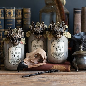 May include: Three glass jars with labels that read "Vampire Dust" and feature a bat design. The jars are sitting on a wooden surface with a skull and an antique key in front of them.