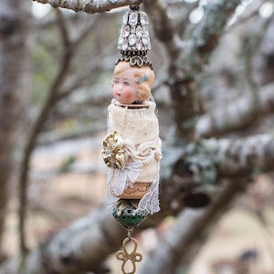 May include: A vintage doll head ornament with a white fabric body, a rhinestone crown, and a green bead hanging from a gold chain.