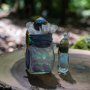 Puede incluir: Dos botellas de vidrio decorativas con purpurina iridiscente en su interior. La botella m&aacute;s grande tiene una tapa de encaje azul y blanco y un adorno dorado. La botella m&aacute;s peque&ntilde;a tiene un adorno verde y dorado. Ambas botellas est&aacute;n sobre una superficie de madera.