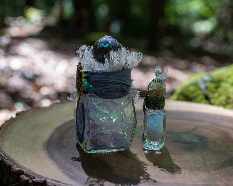 Puede incluir: Dos botellas de vidrio decorativas con purpurina iridiscente en su interior. La botella m&aacute;s grande tiene una tapa de encaje azul y blanco y un adorno dorado. La botella m&aacute;s peque&ntilde;a tiene un adorno verde y dorado. Ambas botellas est&aacute;n sobre una superficie de madera.