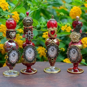 May include: Four decorative glass bottles with red accents and gold-toned metal embellishments. Each bottle features a central oval label with the text Fairy Dust, topped with ornate stoppers including hearts, spheres, and floral metal designs.