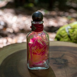 Puede incluir: Una botella de vidrio transparente con un tap&oacute;n de madera oscura y una cinta roja alrededor del cuello. La botella est&aacute; llena de un l&iacute;quido rosa y naranja.
