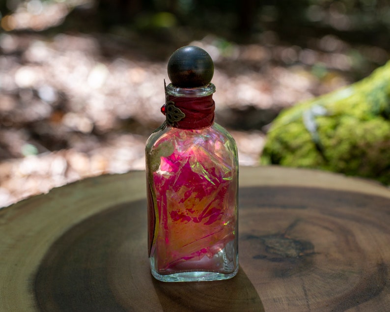 Puede incluir: Una botella de vidrio transparente con un tap&oacute;n de madera oscura y una cinta roja alrededor del cuello. La botella est&aacute; llena de un l&iacute;quido rosa y naranja.